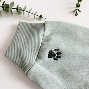 K&ouml;nnte beinhalten: Ein hellgr&uuml;ner Pullover mit einem gestickten schwarzen Pfotenabdruck auf der Vorderseite.