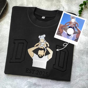 May include: Black sweatshirt featuring an embroidered design of a person with a baby on their shoulders. The word "DAD" is embroidered, along with "EST. 2025". A small photo shows a similar scene.