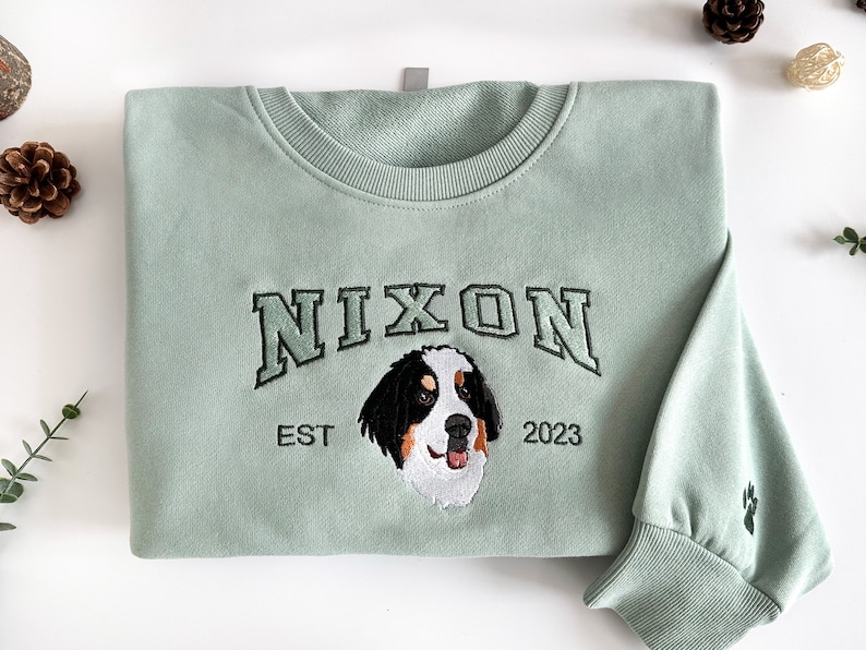 K&ouml;nnte beinhalten: Ein hellgr&uuml;ner Pullover mit einem schwarzen Stickmuster. Das Design zeigt das Wort "NIXON" in einer College-Schriftart, einen Berner Sennenhund und den Text "EST 2023".