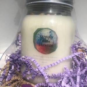 May include: A white candle in a glass jar with a silver lid, wrapped in clear plastic. The candle has a sticker that reads "YAHUAH is the CREATOR." The candle is surrounded by purple and gold shredded paper.