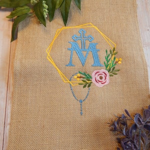 Puede incluir: Un tapiz de pared de tela de arpillera con un diseño bordado de un marco geométrico dorado con una "M" azul y una cruz en su interior. El diseño también incluye un rosario azul y flores rosas y amarillas.