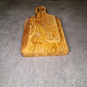 Op de afbeelding: Twee snijplanken van olijfhout. De planken zijn op elkaar gestapeld. De bovenste plank heeft een handvat.