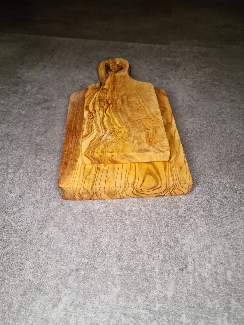 Op de afbeelding: Twee snijplanken van olijfhout. De planken zijn op elkaar gestapeld. De bovenste plank heeft een handvat.
