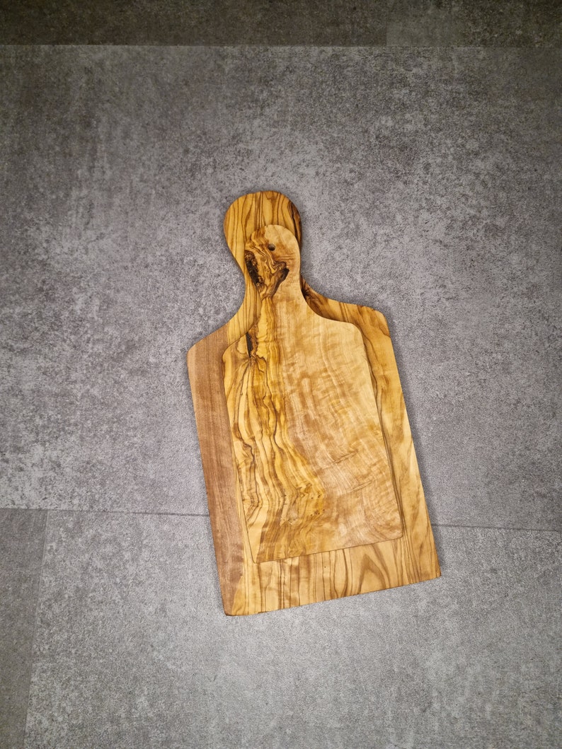 Op de afbeelding: Twee snijplanken van olijfhout. De planken zijn in elkaar geschoven, met de kleinere plank bovenop de grotere plank. De planken hebben een natuurlijke houtnerf en zijn lichtbruin.