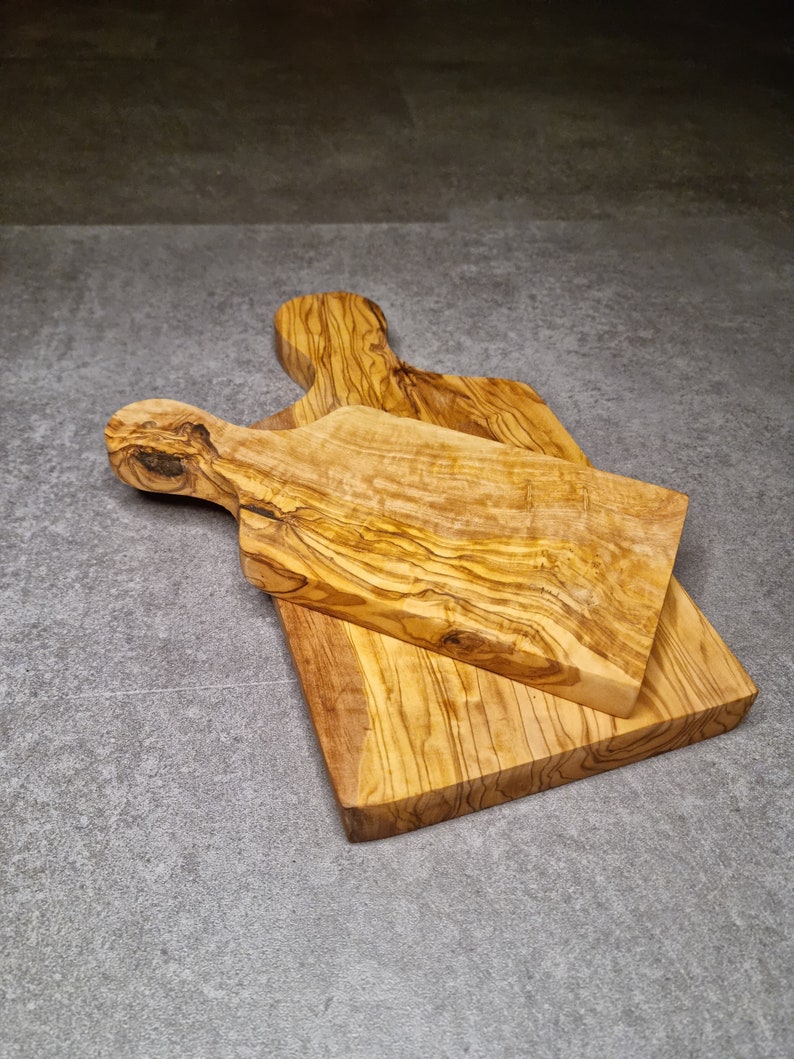 Op de afbeelding: Twee snijplanken van olijfhout, gestapeld op elkaar. De bovenste plank heeft een handvat aan de linkerkant. De planken zijn lichtbruin van kleur met donkerbruine strepen.