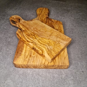 Op de afbeelding: Twee snijplanken van olijfhout. De planken zijn van verschillende grootte en vorm, en beide zijn gemaakt van lichtbruin hout met een natuurlijke nerf.