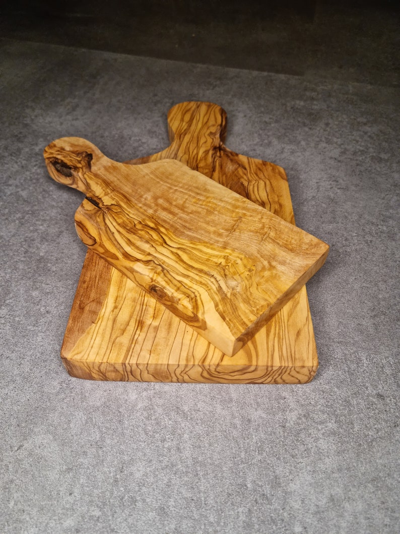 Op de afbeelding: Twee snijplanken van olijfhout. De planken zijn van verschillende grootte en vorm, en beide zijn gemaakt van lichtbruin hout met een natuurlijke nerf.