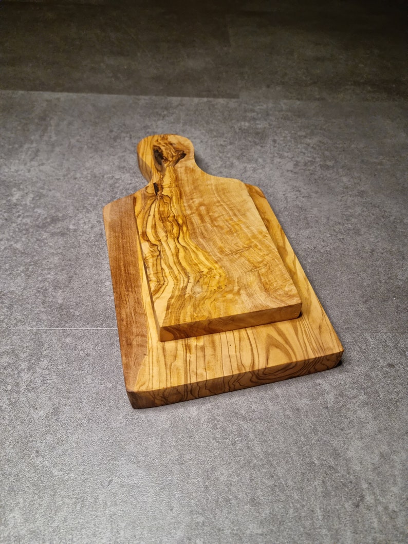 Op de afbeelding: Twee snijplanken van olijfhout. De planken zijn op elkaar gestapeld. De bovenste plank heeft een handvat.