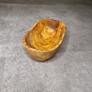 May include: A small, oval-shaped wooden bowl crafted from olive wood. The bowl features a natural wood finish and a light brown hue. The grain of the wood is visible, adding to the rustic appeal of the bowl.