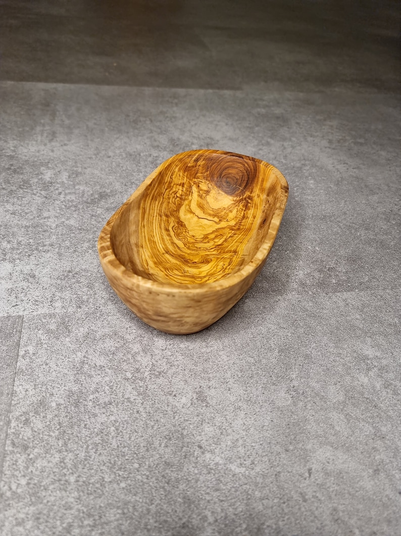 May include: A small, oval-shaped wooden bowl crafted from olive wood. The bowl features a natural wood finish and a light brown hue. The grain of the wood is visible, adding to the rustic appeal of the bowl.
