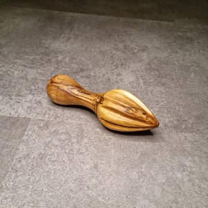 Op de afbeelding: Een houten citruspers gemaakt van olijfhout. De pers heeft een handvat en een puntige ruimer om sap te extraheren. De houtnerf is zichtbaar en toont een natuurlijke, lichtbruine kleur. Ontworpen voor het handmatig persen van citrusvruchten.