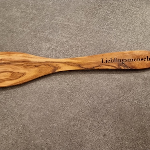 Könnte beinhalten: Ein Holzspatel mit natürlicher Holzmaserung. Der Spatel hat einen gebogenen Griff und die Worte "Lieblingsmensch" sind in den Griff graviert.