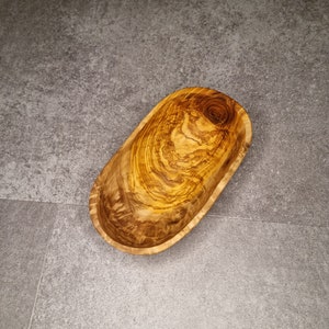 May include: An oval-shaped wooden bowl made from olive wood. The bowl has a natural wood grain pattern.