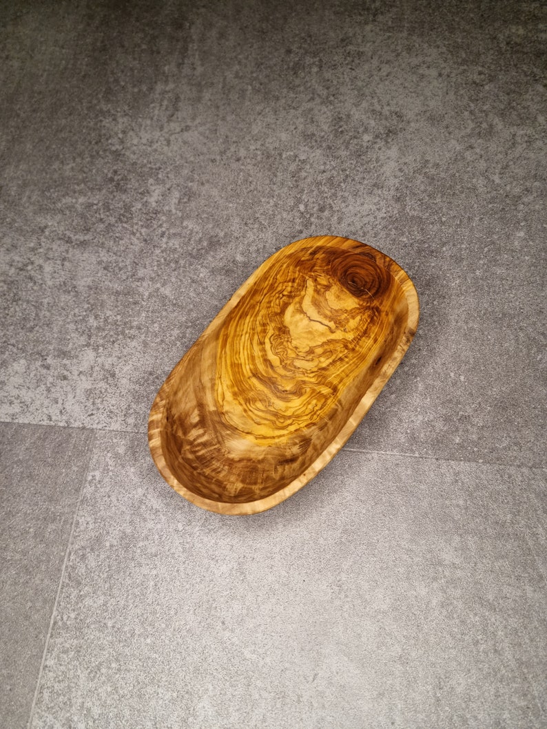 May include: An oval-shaped wooden bowl made from olive wood. The bowl has a natural wood grain pattern.