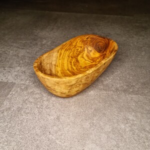 May include: A small, oval-shaped wooden bowl made from olive wood. The bowl has a natural wood grain pattern and is ideal for serving snacks or dips.