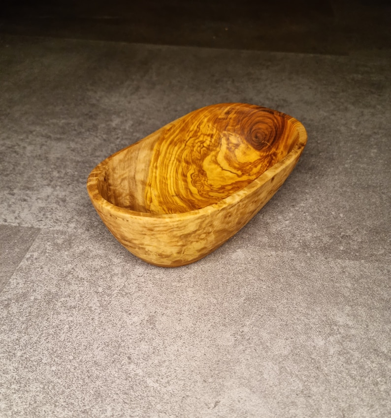 May include: A small, oval-shaped wooden bowl made from olive wood. The bowl has a natural wood grain pattern and is ideal for serving snacks or dips.