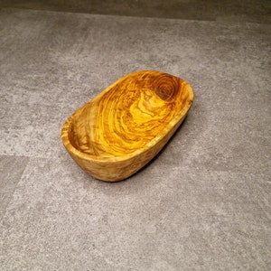 Pode incluir: Uma tigela de madeira pequena, oval, com um padrão natural de grão de madeira. A tigela é feita de madeira de oliveira e tem uma cor castanho claro.