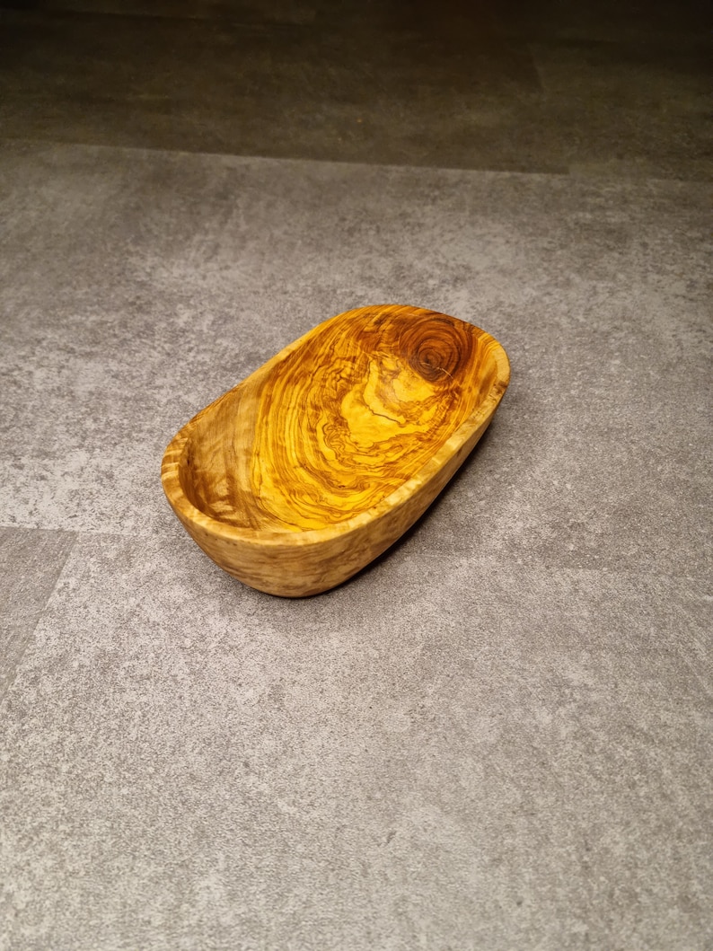 May include: A small, oval-shaped wooden bowl with a natural wood grain pattern. The bowl is made of olive wood and has a light brown colour.