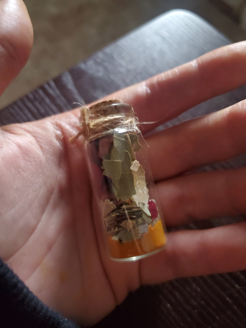 May include: A small, clear glass bottle filled with dried herbs and spices. The bottle is sealed with a cork and twine. The contents include green leaves, yellow powder, and small red petals. The bottle is held in a hand.