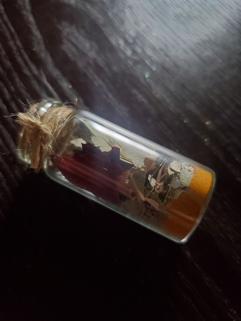 May include: A small, clear glass bottle filled with dried leaves and a layer of orange-colored material. The bottle is sealed with a cork and tied with twine. The background is a dark, textured surface.