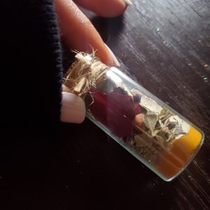 May include: A small, clear glass bottle filled with dried herbs, spices, and a deep red flower. The bottle is sealed with a cork and twine. The contents are visible through the glass, creating a layered effect. The bottle is held against a dark background.