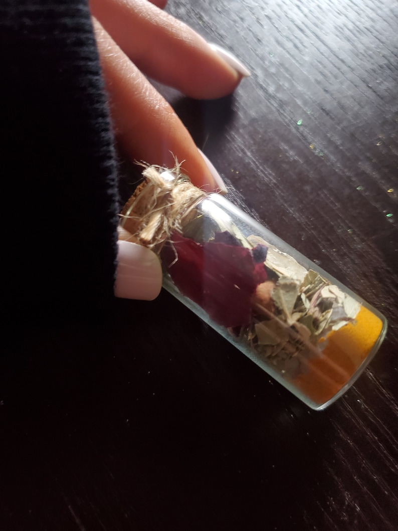 May include: A small, clear glass bottle filled with dried herbs, spices, and a deep red flower. The bottle is sealed with a cork and twine. The contents are visible through the glass, creating a layered effect. The bottle is held against a dark background.