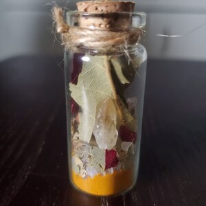 May include: A small, clear glass bottle filled with layers of ingredients, including a vibrant yellow powder, dried leaves, and small crystals. The bottle is sealed with a cork and tied with twine, creating a rustic aesthetic. The contents appear to be a potion or spell.
