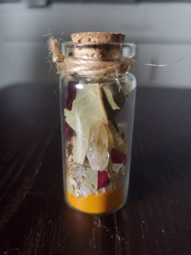 May include: A small, clear glass bottle filled with layers of ingredients, including a vibrant yellow powder, dried leaves, and small crystals. The bottle is sealed with a cork and tied with twine, creating a rustic aesthetic. The contents appear to be a potion or spell.