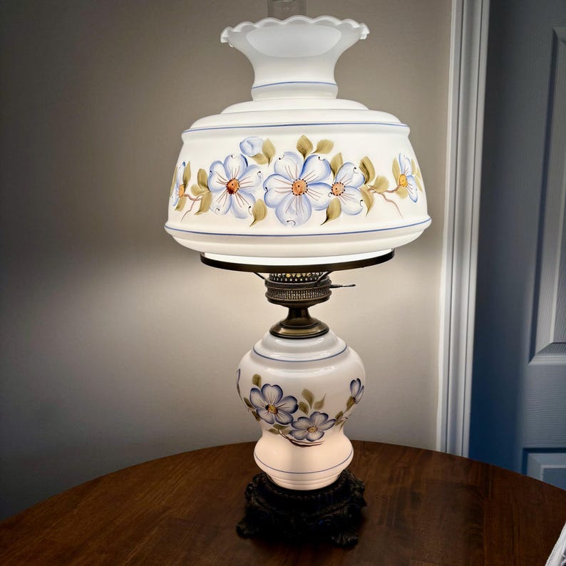 May include: A white table lamp with a floral design on the shade and base. The shade is white with blue and yellow flowers and green leaves. The base is white with blue flowers. The lamp is on a wooden table.