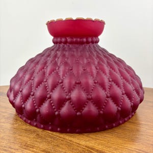 May include: A deep red glass lampshade with a quilted design. The shade has a rounded, bell-like form with a scalloped edge and a smaller, cylindrical top. The surface is textured with raised diamond shapes.