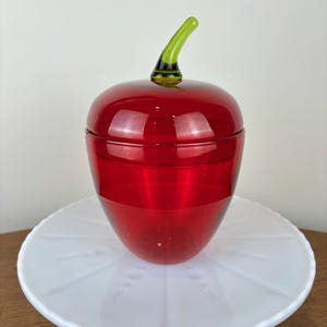Puede incluir: Un tarro de vidrio rojo con forma de manzana con una tapa de vidrio verde en forma de tallo. El tarro está sobre un plato blanco.