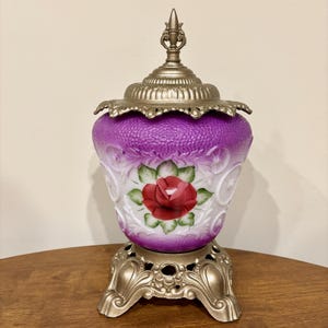 Puede incluir: Lámpara decorativa de estilo vintage con cuerpo de vidrio texturizado morado y base y tapa de metal dorado. La lámpara presenta un diseño de rosa roja pintada a mano sobre fondo blanco. La lámpara mide aproximadamente 30 cm de altura.
