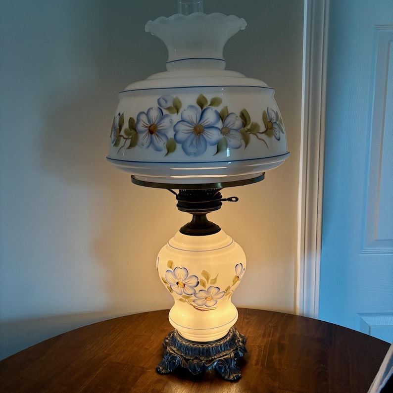 May include: A white glass lamp with a floral design. The lamp has a white shade with blue flowers and green leaves. The base of the lamp is also white with blue flowers and green leaves. The lamp is on a wooden table.