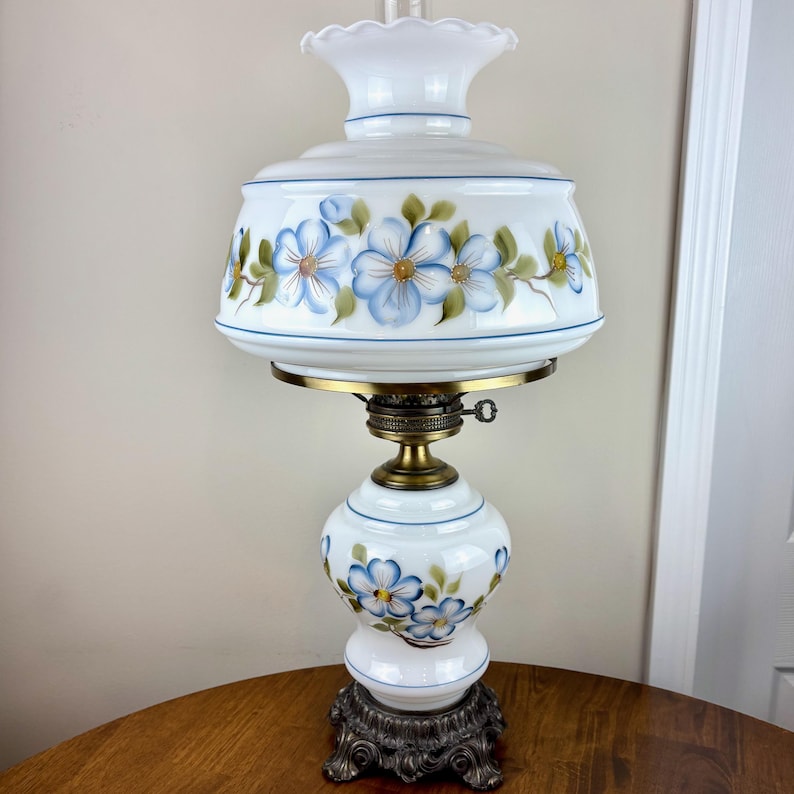 May include: White table lamp with a white glass shade featuring a blue floral design. The lamp has a brass base and a white glass body with a blue floral design.