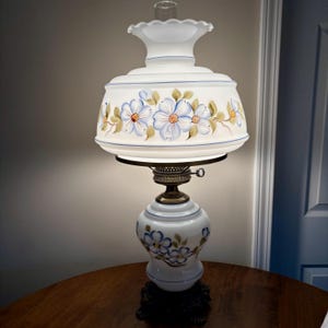 May include: White milk glass lamp with a floral design. The lamp has a white shade with blue and green flowers painted on it. The base of the lamp is also white with blue flowers painted on it.