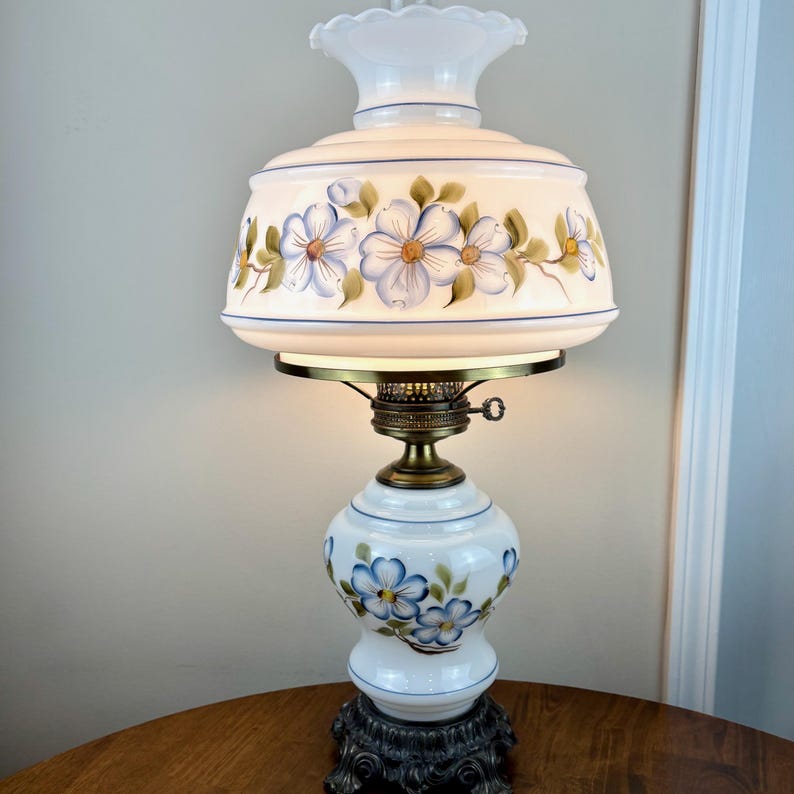 May include: A white and blue floral lamp with a white glass shade and a brass base. The shade has a hand-painted design of blue flowers and green leaves.
