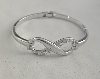 May include: A silver-colored bracelet with an infinity symbol at the center. The infinity symbol is embellished with small, clear gemstones. The bracelet has a hinged clasp and a polished finish, reflecting light. The jewelry is set against a neutral background.