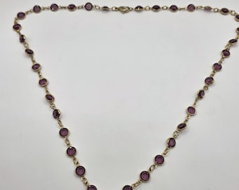 May include: A gold-toned necklace featuring a series of round, deep purple gemstones. The gemstones are evenly spaced along the chain, creating a continuous pattern. The necklace has a simple clasp.