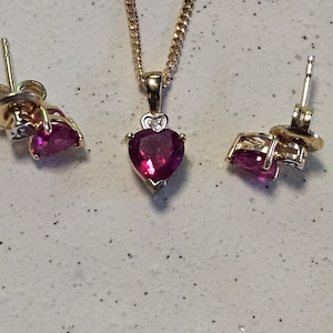 Puede incluir: Un conjunto de joyas en tono dorado con un collar con colgante en forma de corazón y pendientes a juego. El colgante y los pendientes muestran una piedra preciosa rosa intenso en forma de corazón. El conjunto se exhibe sobre una superficie clara.