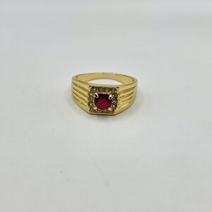 May include: A gold ring featuring a square, red gemstone, encircled by smaller clear stones. The ring's band has a textured pattern. The ring is displayed on a white backdrop.