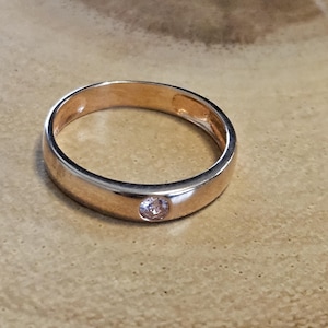 Puede incluir: Un anillo de banda de oro simple con una sola piedra pequeña y clara engastada en el centro.