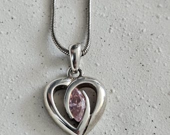 Peut inclure: Pendentif argenté en forme de cœur avec une pierre précieuse rose au centre, suspendu à une chaîne argentée. Le pendentif présente un motif de cœur ouvert avec une finition polie. La pierre précieuse est de forme ovale. Le collier est présenté sur une surface gris clair.