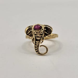 Peut inclure: Une bague dorée représentant un éléphant. La tête de l'éléphant est ornée d'une petite pierre précieuse rouge ovale. La monture de la bague et les détails de l'éléphant présentent des motifs texturés.