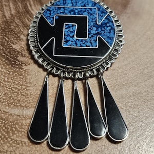 Puede incluir: Un colgante de plata con un diseño geométrico negro y azul y una franja de acentos negros en forma de lágrima.