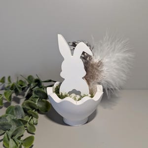 Könnte beinhalten: Eine weiße Ostersilhouette eines Hasen sitzt in einem weißen, eiförmigen Behälter, der mit grünem Moos und Federn gefüllt ist. Der Hase ist der Mittelpunkt, mit sichtbaren Ohren und Schwanz. Das dekorative Stück ist vor einem neutralen Hintergrund platziert.