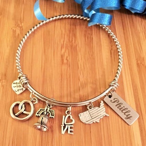 Op de afbeelding: Zilveren armband met meerdere bedels, waaronder een pretzel, de Liberty Bell, een hart met "LOVE" erop, een kaart van de VS en een tag met "Philly" erop.
