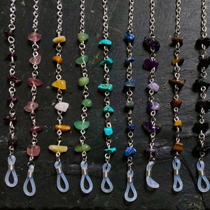 May include: A selection of spectacle chains featuring silver-tone chains and vibrant gemstone beads. Each chain showcases a unique mix of stones, such as pink, yellow, green, blue, and purple. The chains terminate with clear rubber loops for securing to glasses.