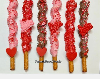 May include: Six chocolate-covered pretzel rods decorated with pink, red, and brown chocolate and sprinkles. The pretzels are topped with red heart-shaped chocolates and the word "Love" in red.