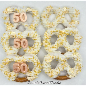 May include: Six white chocolate-covered pretzels decorated with gold sprinkles and edible gold leaf. Each pretzel has a rose gold number "50" on top, perfect for a milestone birthday celebration. The image is branded with "Pamela Sweet Treats".