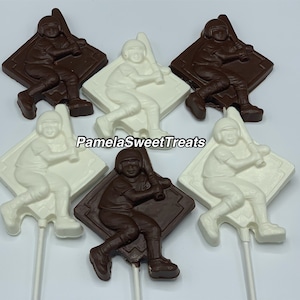 May include: Six chocolate baseball player lollipops. Three are white chocolate and three are dark chocolate. Each lollipop features a baseball player in a batting stance holding a bat. The lollipops are on white sticks.